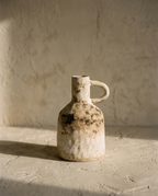 Single-Handle Pottery Vase in mottled cream and brown coarse ceramic, on a plaster ledge in warm raking light