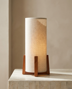 Linen Drum Table Lamp with a cylindrical linen shade on a wooden cross base, glowing warm on a pale stone surface