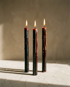 Black taper candles with red wax running down them, lit and standing on a plaster surface