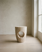 A travertine-look drum side table with a circular cut-out through one side, on pale oak flooring