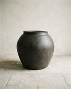 A round-bellied planter pot in a dark aged brown finish, standing on a pale stone floor