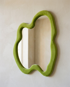 An irregular wavy wall mirror in a green velvet-wrapped frame, mounted on a bone plaster wall