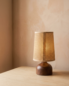 A cordless table lamp with a woven jute shade on a turned dark wood dome base, glowing warm on an oak surface