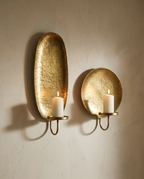 A pair of hammered brass wall sconces, one oval and one round, each holding a lit pillar candle