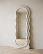 A full-length floor mirror in a wavy scalloped fabric-wrapped frame, leaning against a plaster wall