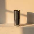 Mirror-polished steel cylinder vase on a plaster ledge in golden hour light